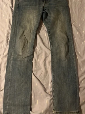 Blå raka jeans i klassisk modell - Säljer ett par blå jeans med rak passform och klassisk look. Jeansen har en ljus tvätt och subtila slitningar framtill för en avslappnad stil. Perfekta till vardags och lätta att matcha med olika outfits.