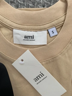 Beige t-shirt från Ami Paris - Beige t-shirt från Ami Paris med klassisk rund hals och en liten röd broderad logga med hjärtdetalj på bröstet. Tillverkad i mjuk bomull och har en enkel, clean design som passar till allt.