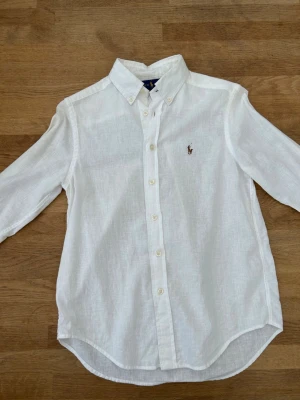 Vit linneskjorta från Ralph Lauren - Klassisk vit skjorta från Ralph Lauren i svalt linne. Skjortan har lång ärm, button-down krage och den ikoniska broderade loggan på bröstet. Perfekt till både jeans och chinos för en clean och stilren look. Märke finns på ena axel oklart om den går bort