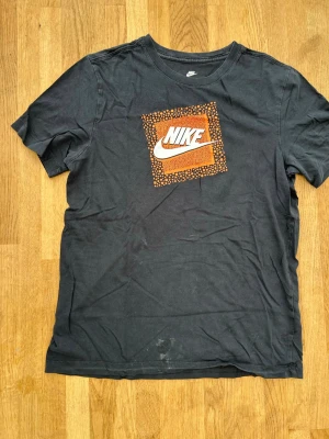 Svart Nike t-shirt med logga - Svart t-shirt från Nike med klassisk logga i vitt och orange på bröstet. T-shirten har rund halsringning och korta ärmar. Tillverkad i mjuk bomull för en bekväm känsla. Perfekt för en avslappnad och sportig stil. Märke finns men borde gå bort i tvätten 