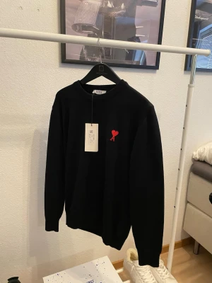 Ami Paris  - Säljer en exklusiv och stilren AMI Paris stickad tröja i klassisk svart med den ikoniska röda Ami de Cœur-loggan broderad på bröstet. En tidlös premiumtröja som kombinerar minimalistisk design med högsta kvalitet och passar perfekt till både vardag och mer uppklädda tillfällen.  ✔️ Helt ny – aldrig använd ✔️ Originaltags sitter kvar ✔️ 100% äkta ✔️ Perfekt skick – som direkt från butik ✔️ Otroligt mjuk och exklusiv känsla  En lyxig garderobsklassiker från ett av världens mest eftertraktade modemärken