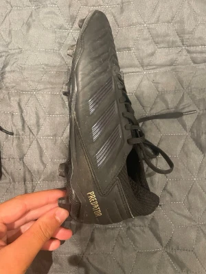 Adidas Predator svarta fotbollsskor - Säljer ett par svarta Adidas Predator fotbollsskor med klassisk design och snörning. Skorna har mönstrad ovandel i syntetmaterial, fasta dobbar för spel på gräs och diskreta detaljer i guld. Perfekta för dig som vill ha kontroll och stil på planen.