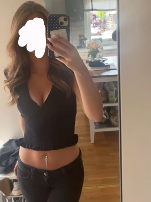 Svart croppad v-ringad topp - Snygg svart croppad topp med v-ringning och volangkant nertill. Toppen är ärmlös och har en figurnära passform som framhäver midjan. Perfekt för dig som gillar en trendig och stilren look.