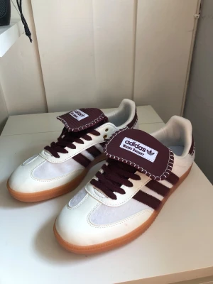 Adidas Wales Bonner sneakers vinröd/vit - Säljer ett par Adidas Wales Bonner sneakers i vitt och vinrött med klassiska tre ränder på sidan, grov sula i gummi och unika detaljer på plösen. Skorna har låga skaft och är tillverkade i en mix av läder och textil. Perfekta för dig som gillar retrovibbar och cool design.