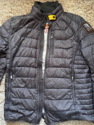 Grå UGO  pufferjacka från Parajumpers - Snygg grå pufferjacka från Parajumpers med gul detalj vid halsen och patch på ärmen. Jackan har dragkedja framtill, två sidofickor med dragkedja och en bröstficka. Tillverkad i lätt, quiltat material som håller värmen under kalla dagar. 
