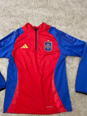 Spansk landslagströja Adidas röd/blå - Snygg långärmad träningströja från Adidas för det spanska landslaget. Tröjan är röd med blå ärmar och sidor, har halv dragkedja och landslagets emblem på bröstet. Tillverkad i funktionsmaterial med Aeroready-teknologi. Perfekt för fotbollsträning.