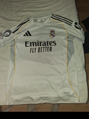Real Madrid matchtröja Mbappé - Säljer en vit Real Madrid matchtröja från Adidas med svarta detaljer och gula linjer. Tröjan har klubbmärke, sponsortryck och nummer 9 med Mbappé på ryggen. Tillverkad i lätt och ventilerande material, perfekt för fotbollsträning eller match.