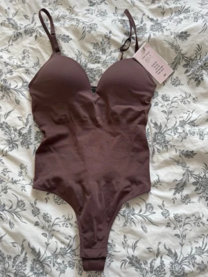 Brun shaping-body från H&M - Snygg brun shaping-body från H&M med vadderade kupor och justerbara axelband. Modellen har en smickrande passform och är tillverkad i ett mjukt, stretchigt material som ger lätt formande effekt. Endast testad med kläder imellan!!⭐️