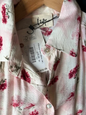 Blommig ärmlös klänning från Isay - Söt ärmlös klänning från Isay i ljusrosa nyans med blommigt mönster i rött och grönt. Klänningen har krage, knappar framtill och ett bälte i midjan för en snygg siluett. Perfekt för varma dagar och somriga tillfällen. Helt ny, aldrig använd.