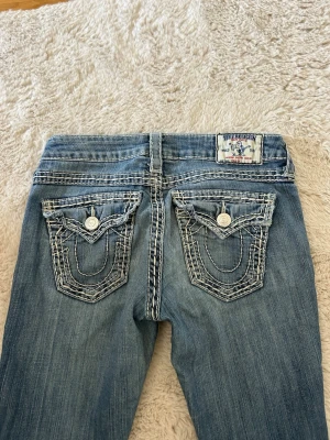 True Religion jeans - Säljer ett par blå jeans från True Religion med ikoniska kontrastsömmar och snygga fickdetaljer bak. Klassisk patch med logga på linningen och silverfärgade knappar. Jeansen har bootcut passform och är i denim.