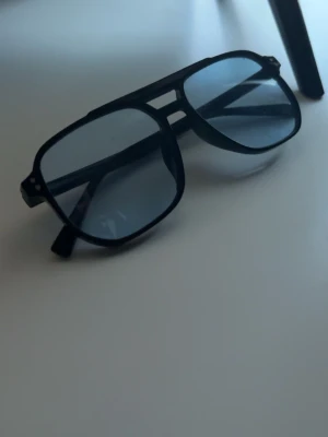 Svarta pilotbågar solglasögon - Snygga solglasögon med svarta pilotbågar och mörka glas. Klassisk aviator-modell med dubbla bryggor och diskreta silvriga detaljer vid gångjärnen. Perfekta för dig som vill ha en stilren och tidlös look.