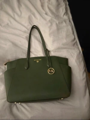 Grön handväska från Michael Kors - Stilren grön handväska från Michael Kors i slätt läder med guldfärgade detaljer och MK-hänge. Väskan har två långa handtag, dragkedja upptill och ett rymligt, quiltat innerfack med extra fickor. Perfekt för dig som vill ha en snygg och praktisk väska.