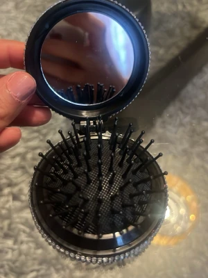 Glittrig hopfällbar hårborste med spegel - Säljer en svart hopfällbar hårborste med inbyggd spegel. Utsidan är täckt av silvriga strass-stenar som ger ett riktigt glittrigt och lyxigt intryck. Perfekt att ha i väskan för snabba touch-ups. Borsten har mjuka piggar och är enkel att fälla ihop.