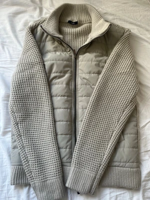 Ljusgrå stickad cardigan med dragkedja - Snygg ljusgrå cardiganjacka med stickade ärmar och rygg samt vadderad framsida. Jackan har hög krage, hel dragkedja och ribbade muddar. Perfekt för lager på lager och ger en modern look. Köpt i Rom, Italien i en affär med autentiska italienska plagg och stiler. Knappt använd!