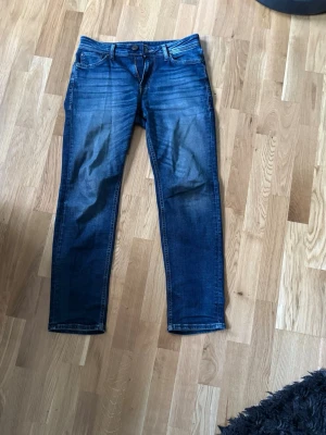 Mörkblå raka jeans med slitningar - Säljer ett par mörkblå jeans med raka ben och snygga slitningar framtill. Jeansen har klassisk femficksdesign, knappgylf och kontrastsömmar. Perfekta till en avslappnad stil och passar till det mesta. Materialet är troligtvis bomull med lite stretch.
