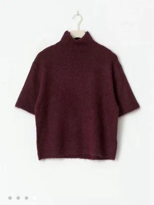 Vinröd stickad tröja med halv polo - Mysig vinröd stickad tröja med halv polokrage och korta ärmar. Tröjan har en boxig passform och är tillverkad i ett mjukt, luddigt material som ger en avslappnad look. Perfekt att styla med jeans eller kjol för en trendig outfit.