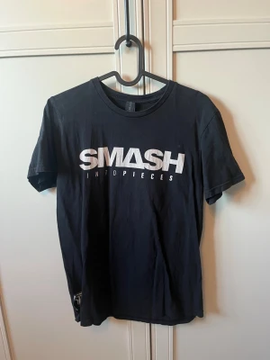 Svart Smash Into Pieces t-shirt - Cool svart t-shirt från Smash Into Pieces Clothing med vit logga och text på bröstet. T-shirten har korta ärmar och en patch med bandets logga och detaljer på sidan. Tillverkad i mjuk bomull för skön känsla. Säljer också en till likadan i samma storlek!