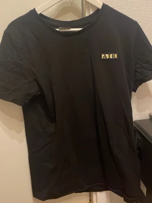 Svart AIK t-shirt med tryck - Säljer en svart t-shirt från AIK med klubbens lilla logga på bröstet och stort tryck '1891' i vitt och gult på ryggen. Klassisk passform och rund halsringning. Perfekt för dig som vill visa ditt AIK-stöd.
