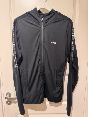 Svart hoodie från BOSS med logga - Snygg svart hoodie från BOSS med dragkedja framtill och vita BOSS-loggor längs ärmarna och på bröstet. Tillverkad i ett mjukt och stretchigt material som känns skönt mot huden. Huva med logga på kanten och två fickor framtill. Perfekt för en chill och sportig look.