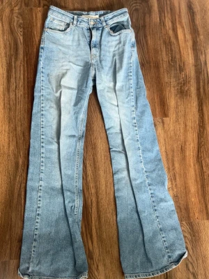 Ljusblå bootcut jeans - Säljer ett par ljusblå bootcut jeans med klassisk femficksdesign och hög midja. Jeansen har en snygg tvättad look och vida ben som ger en retrovibe. Tillverkade i denim, perfekta för dig som gillar avslappnad men trendig stil.