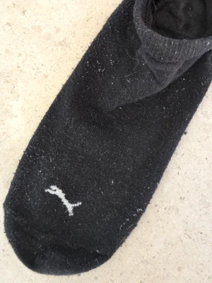 Svart ankelstrumpa från Puma - Svart ankelstrumpa från Puma med diskret logga i vitt framtill. Tillverkad i mjuk bomullsblandning som sitter skönt på foten. Perfekt för sneakers och låga skor.