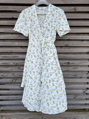 Vit blommig omlottklänning NA-KD - Lång, vit omlottklänning från NA-KD med blommigt mönster i blått och gult. Klänningen har korta ärmar, volangdetaljer och knytband i midjan. Perfekt för sommarens alla tillfällen och ger en romantisk vibe. 