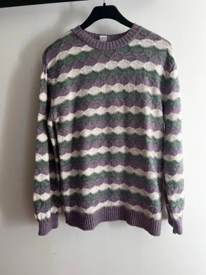 Mönstrad stickad tröja från Lindex - Stickad tröja från Lindex med vågmönster i lila, grönt och vitt. Tröjan har rund halsringning, ribbade muddar och är gjord i mjukt material. Perfekt för dig som gillar retrovibbar och färgkombinationer. Aldrig använd