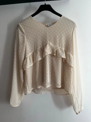 Krämvit blus med volang från Lindex - Söt krämvit blus från Lindex med diskret prickigt mönster och volangdetalj framtill. Blusen har lång ärm och lätt transparent tyg som ger en luftig känsla. Perfekt att styla med jeans eller kjol för en trendig look.