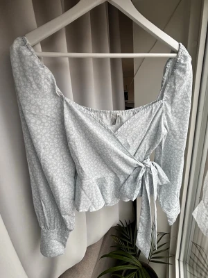 Ljusblå omlottblus med volang - Supersöt ljusblå omlottblus med småblommigt mönster och volangkant nedtill. Blusen har långa ärmar med breda muddar och knyts i sidan för en snygg passform. Perfekt till jeans eller kjol för en trendig look.