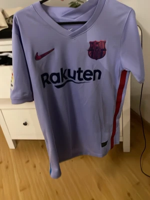 Ljuslila FC Barcelona matchtröja - Säljer en ljuslila FC Barcelona matchtröja från Nike med klubbmärke och Rakuten-logga på bröstet. Tröjan har korta ärmar, V-ringning och röda samt blå detaljer på sidorna. Materialet är lätt och andas, perfekt för fotboll eller träning.