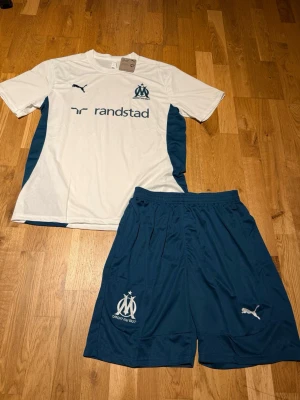 Marseille summer set white/blue - Säljer ett Marseille matchställ från Puma med vit t-shirt och blå shorts. Tröjan har klubbmärke och sponsortryck framtill samt blå sidopaneler. Shortsen är blå med klubbmärke och Puma-logga. Perfekt för fotbollsträning eller match.