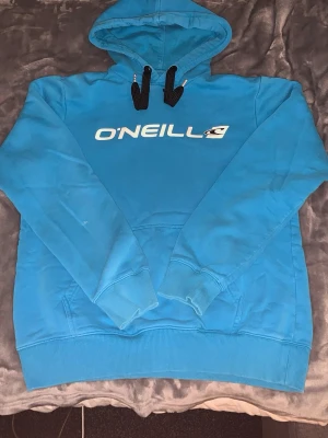 Blå hoodie från O'Neill - Snygg blå hoodie från O'Neill med vit logga på bröstet och svartvita dragsnören i huvan. Klassisk känguruficka framtill och ribbade muddar vid ärmar och nederkant. Perfekt för en avslappnad streetwear-look.