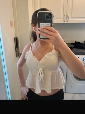 Vit volang topp med knyt och smock - Söt vit linnetopp med volangdetaljer och smockad överdel. Toppen har smala axelband, v-ringning och knyt framtill. Perfekt för sommaren och ger en luftig känsla. Passar till både jeans och kjol.