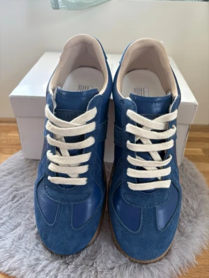 Maison Margiel sneakers blå mocka - Snygga blå sneakers med ovandel i mocka och släta partier i läder. Skorna har vita snören och en klassisk gummisula med mönster undertill. Insidan är ljus och skorna har en retroinspirerad look som passar till många stilar.