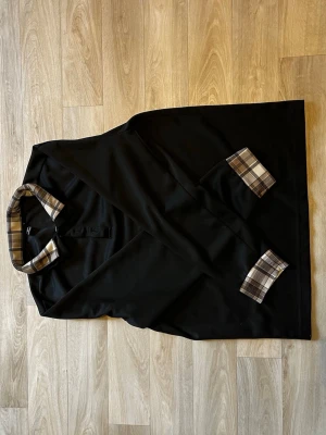 Svart långärmad skjorta med rutiga detaljer - Svart långärmad skjorta med rutigt mönster på krage och ärmslut. Skjortan har knappar framtill och en klassisk passform. Perfekt för dig som gillar stilrena plagg med en twist.