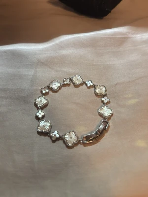 Silverfärgat armband med blommönster - Snyggt armband i silverfärg med fyrklöverformade länkar och blommönster på varje länk. Armbandet har en dekorativ och unik design med små detaljer mellan länkarna. Perfekt för dig som gillar stilrena accessoarer med en twist.