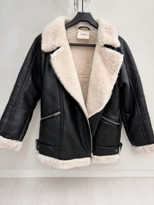 Svart skinnjacka med fuskpäls från Pull&Bear - Stilren svart skinnjacka från Pull&Bear med mjukt beige fuskpälsfoder. Jackan har bred krage, dragkedja i sned modell, två snedställda fickor med dragkedja och spänndetaljer vid midjan. Perfekt för dig som vill ha en trendig och varm look.