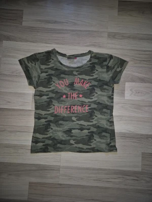 Camo t-shirt med texttryck - Säljer en grön camouflagemönstrad t-shirt med korta ärmar och rund hals. På bröstet står det 'YOU MAKE THE DIFFERENCE' i rosa text. Perfekt för dig som gillar streetstyle och vill sticka ut lite extra.