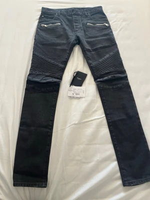 Svarta bikerjeans från Balmain - Säljer ett par helt oanvända svarta bikerjeans från Balmain med coola ribbade detaljer över knäna och dragkedjor vid fickorna. Slim fit-modell som ger en snygg siluett. Perfekta för dig som vill ha en edgy och trendig look.