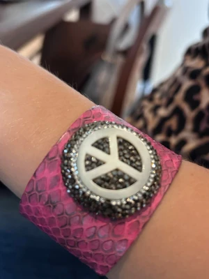 Brett rosa armband med peace-märke - Coolt brett armband i rosa skinn och ett stort peace-märke med glittriga strasstenar, ger cool bling-effekt. Armbandet är 4 cm brett och är reglerbart så det passar olika handleder. Perfekt för dig som vill sticka ut och addera lite edge till din stil. Unikt och one-of-a-kind.