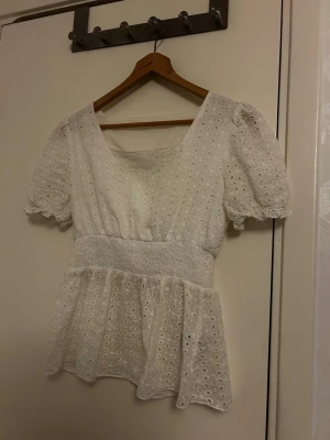 Vit blus med broderade detaljer - Söt vit blus med broderade hålmönster och puffiga korta ärmar. Modellen har en bred smock i midjan som ger en markerad siluett och volang nedtill. Perfekt för dig som gillar romantisk och feminin stil. 