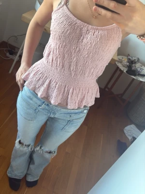 Ljusrosa linne storlek xs Gina trico  - Söt ljusrosa linnetopp med smockad struktur och volang nedtill. Toppen har tunna axelband  Perfekt för sommaren de varma dagar och enkel att styla med jeans eller kjol.