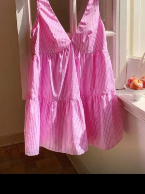 Rosa randig kortklänning - Söt rosa kortklänning med tunna vita ränder och v-ringad hals. Klänningen har en luftig passform och är ärmlös, perfekt för varma dagar. Tillverkad i ett lätt bomullstyg som känns skönt mot huden.