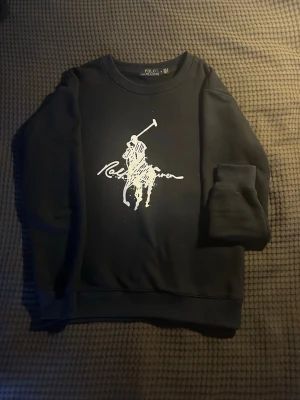 Svart sweatshirt från Polo Ralph Lauren - Svart sweatshirt från Polo Ralph Lauren med stor vit logga och polospelare på bröstet. Klassisk rund halsringning och ribbade muddar vid ärmslut och nederkant. Tillverkad i mjuk bomullsblandning för skön passform. Storlek M men passar både S och M. Knappast använd som ny.