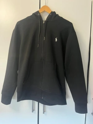 Svart hoodie från Polo Ralph Lauren - Svart hoodie med dragkedja från Polo Ralph Lauren. Klassisk design med vit broderad logga på bröstet, huva med dragsko och två fickor framtill. Mjuk bomullsblandning som är skön att bära. Perfekt för en avslappnad och stilren look.