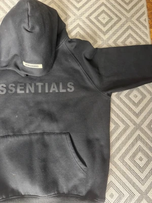 Svart Essentials hoodie med tryck - Säljer en svart hoodie från Essentials med stor logga tryckt över bröstet och en mindre logga på huvan. Hoodien har klassisk känguruficka framtill och är gjord i mjukt, tjockt material. Perfekt för dig som gillar streetwear och enkel stil.