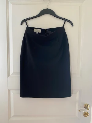 Svart kortkjol från Jennifer Collection - Stilren svart kortkjol från Jennifer Collection. Kjolen har en enkel, rak passform och tunna axelband upptill. Perfekt att matcha med en crop top eller stickad tröja. Klassisk design som funkar till många olika looks.