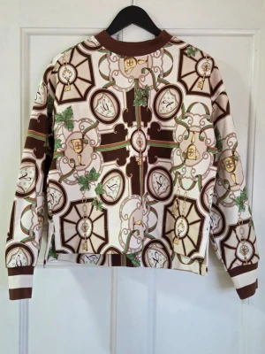 GANT American Luxe C-Neck Sweat - Helt ny och aldrig använd. Unik sweatshirt från Gant med brunt, beige och grönt mönster av klockor och kedjor. Tröjan har ribbade muddar och krage i brunt och vitt. Perfekt för dig som gillar statement-plagg och coola retrovibbar.