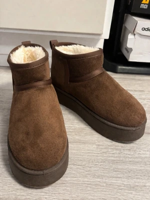 Bruna fodrade stövletter i mocka-look - Mysiga bruna stövletter med tjock platåsula och mjukt, vitt foder på insidan. Utsidan är i mocka-liknande material och skon har en enkel, stilren design med dragflik bak. Perfekta för kalla dagar när du vill hålla värmen och ändå se snygg ut.