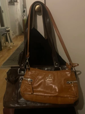 Brun handväska från Adax i skinn - Snygg brun handväska från Adax i äkta skinn med två framfickor med dragkedja och justerbar axelrem. Väskan har silverfärgade detaljer och en stilren, rektangulär form som passar perfekt till en trendig vardagslook.